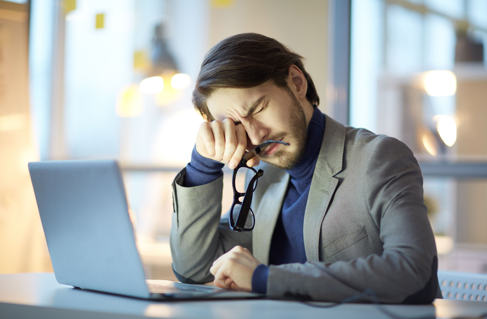 A man sitting at a desk in an office with his laptop and holding his glasses in his right hand as he rubs his eyes