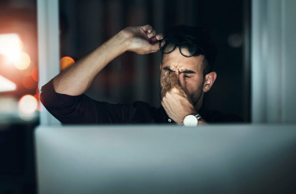 A tired person working at a computer removes their glasses and rubs their eyes.